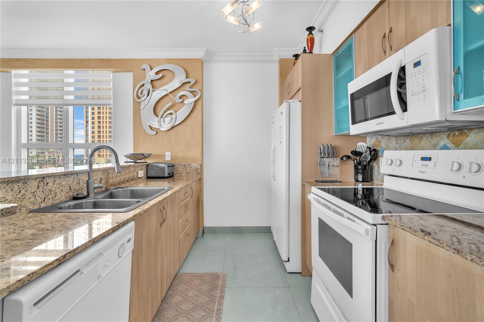 Kitchen featuring light wood-finish cabinetry, stone-surface countertops, dual-basin stainless steel sink, tile flooring, and integrated white appliances