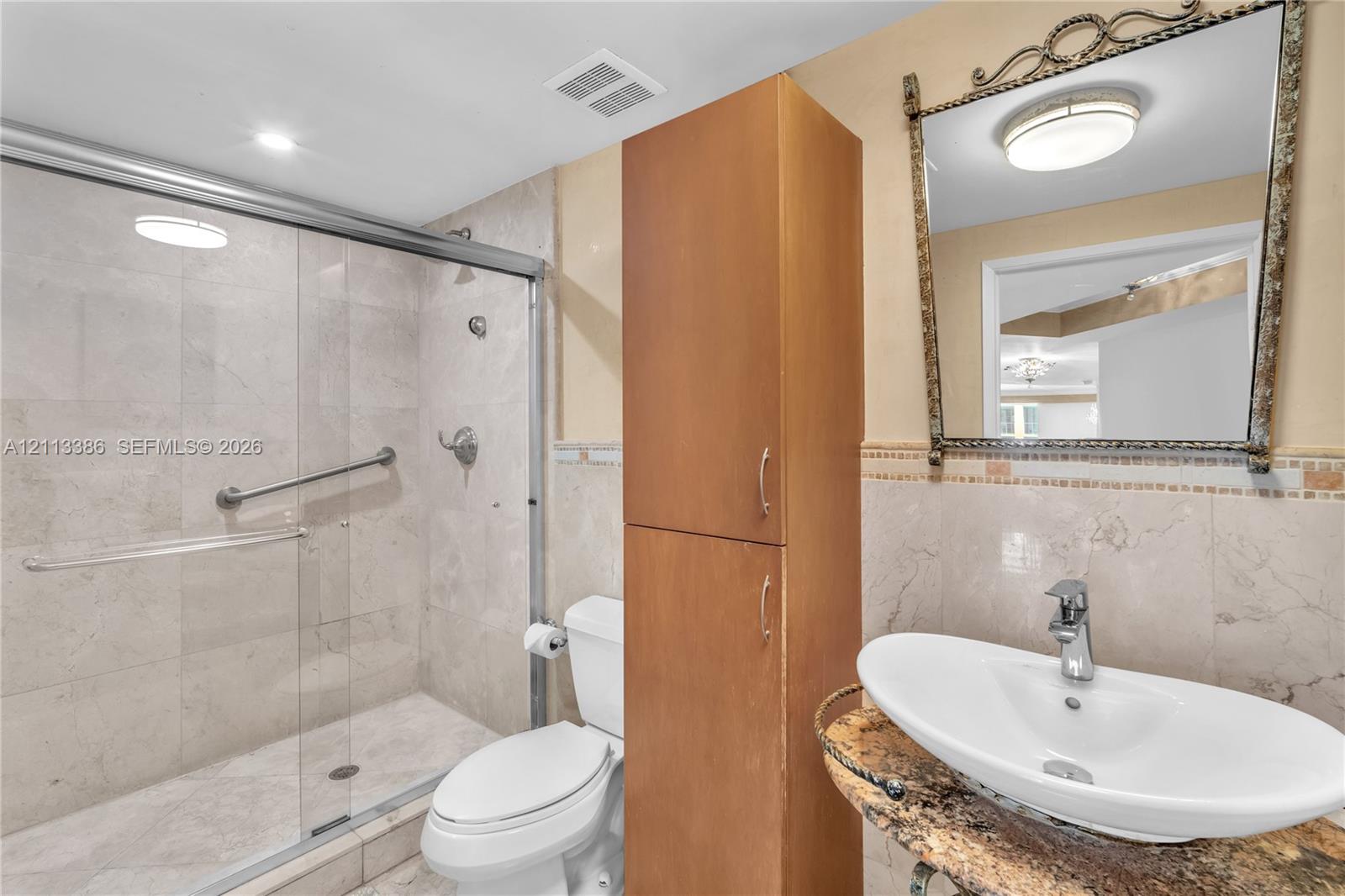 Walk-in shower with sliding glass enclosure and tiled walls, stone-finish countertop with vessel sink, tall built-in wooden cabinetry, and decorative wall mirror
