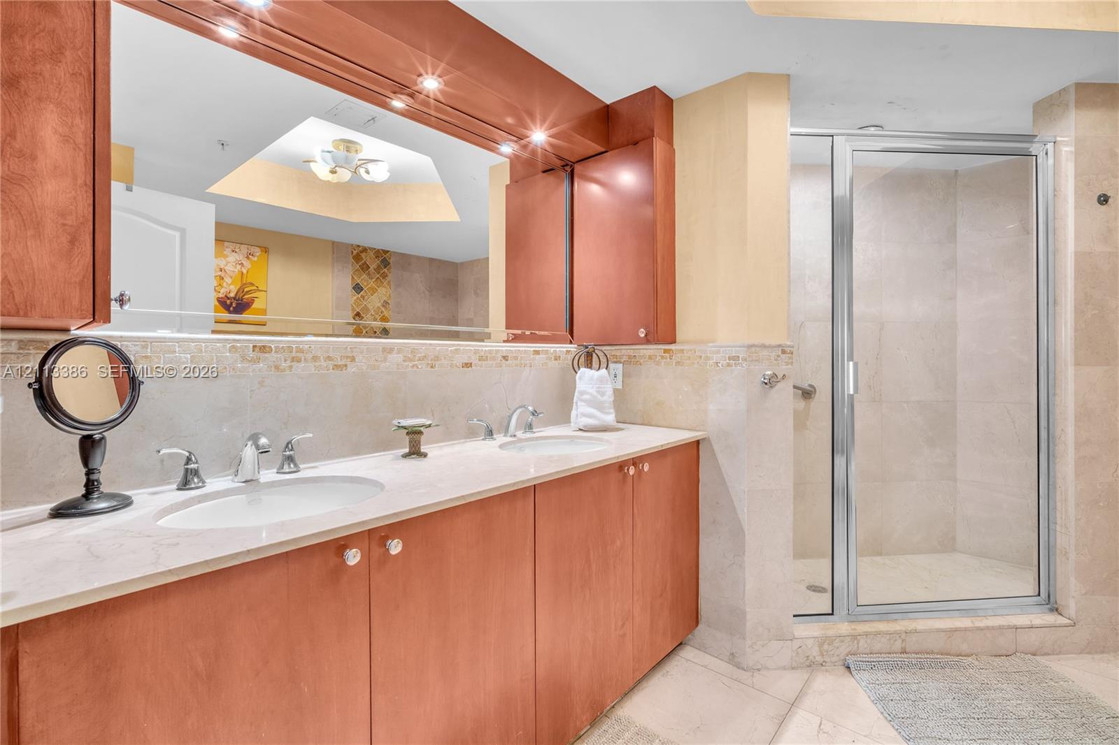 Dual-sink vanity with stone countertop, wood cabinetry, large recessed mirror, and glass-enclosed shower with tiled surround