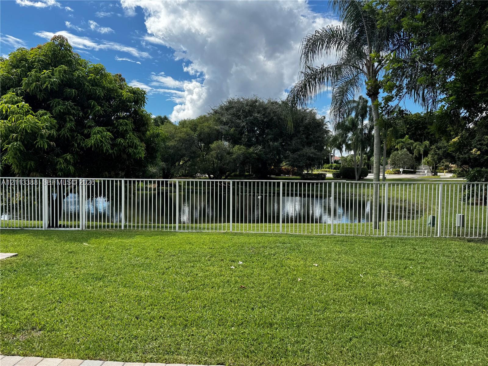 Outdoor space featuring a manicured lawn, white metal fencing, mature trees, and a pond