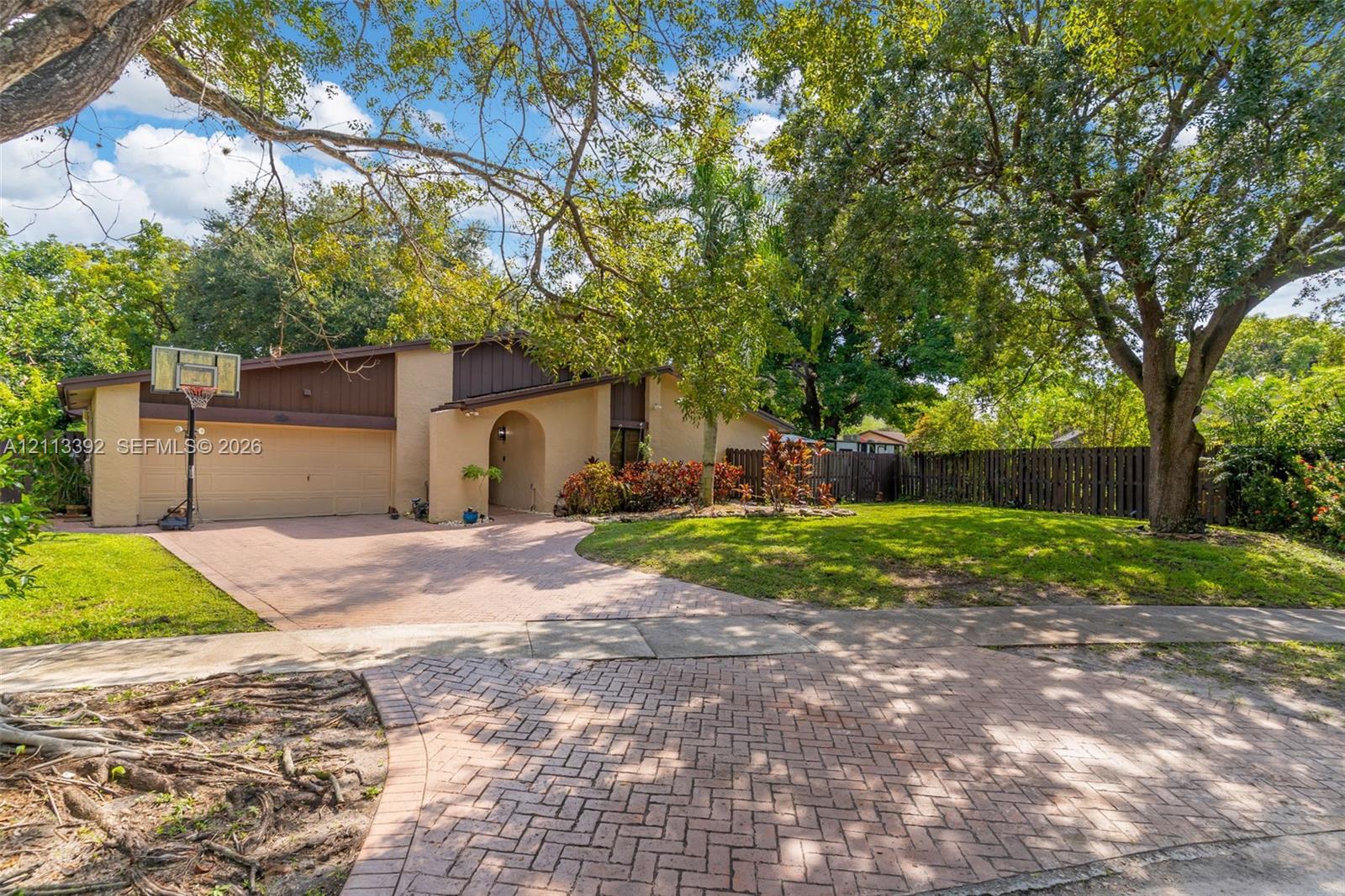 Exterior elevation featuring a multi-tonal facade, paved brick driveway, attached two-car garage, mature landscaping, and fenced perimeter