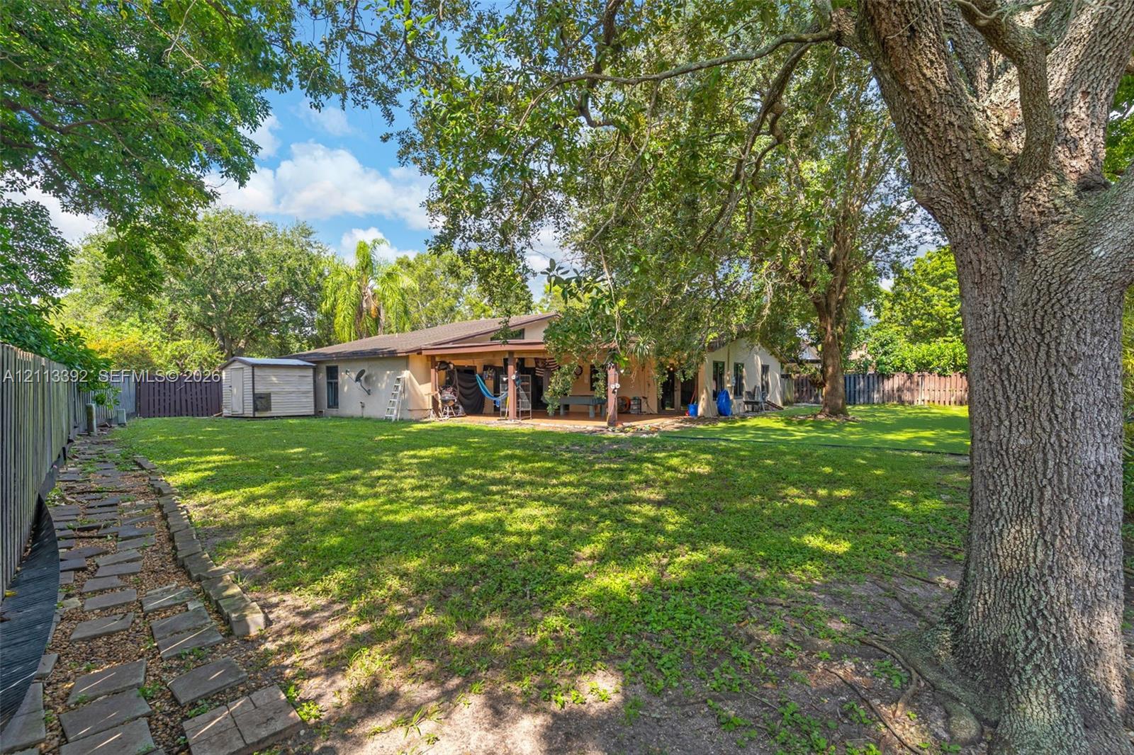 Expansive backyard featuring a covered patio, mature canopy trees, a grass lawn, and stone paver walkway