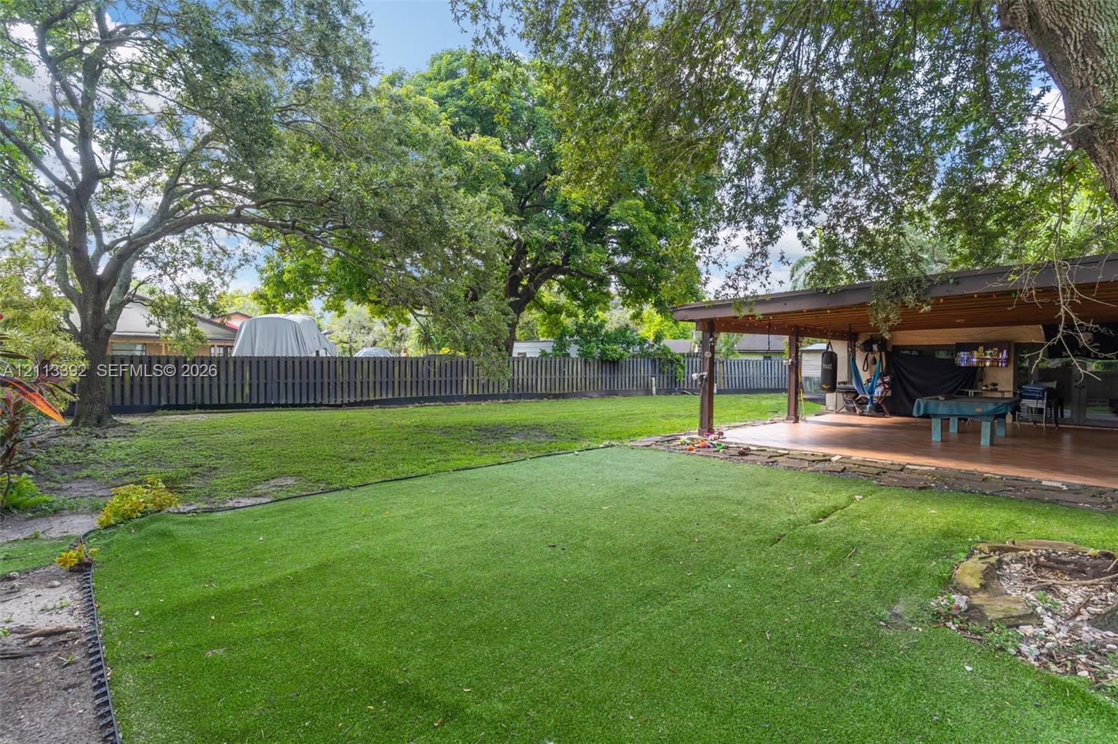 Expansive backyard featuring a covered patio, synthetic turf lawn, perimeter wood fencing, and mature canopy trees