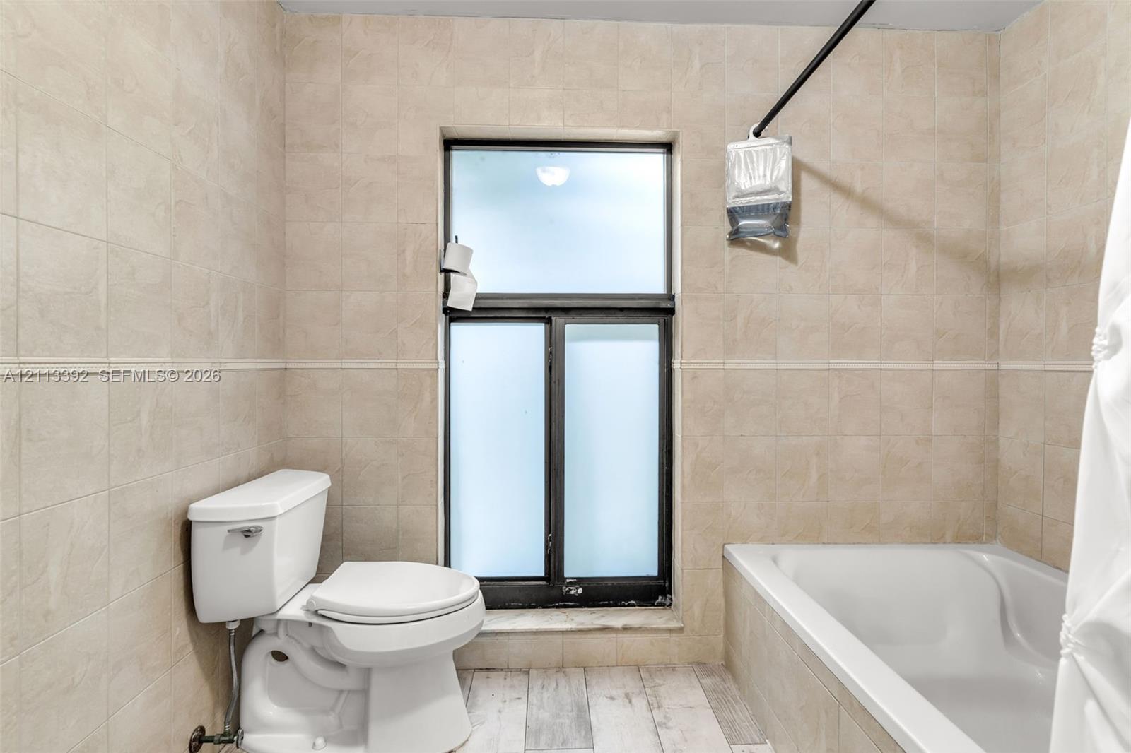 Bathroom featuring neutral tile wall cladding, wood-finish flooring, a white soaking tub, and a window with frosted glass panes