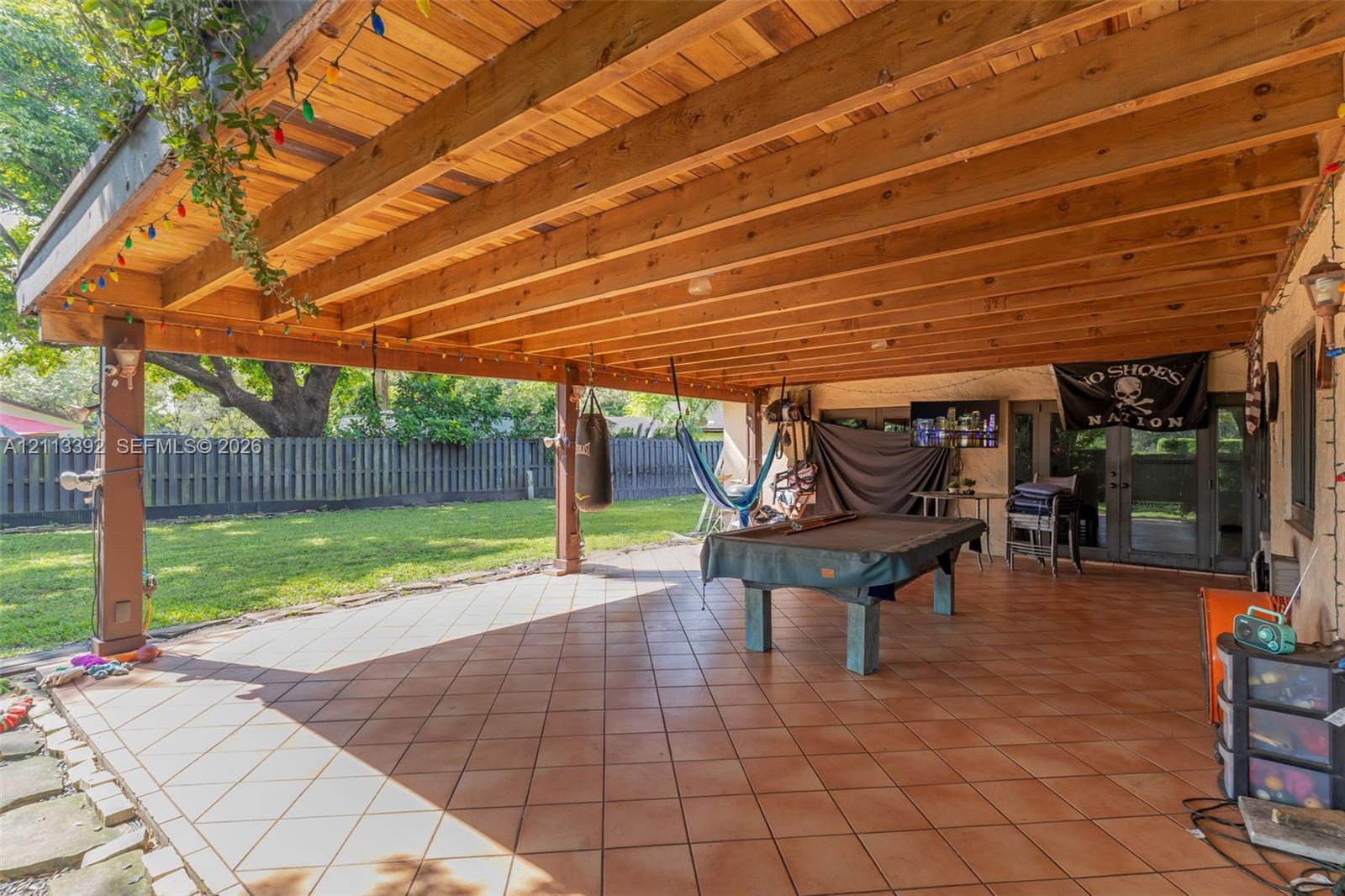 Covered patio featuring a timber-frame ceiling, tile flooring, and vertical support posts, overlooking a lawn enclosed by a privacy fence