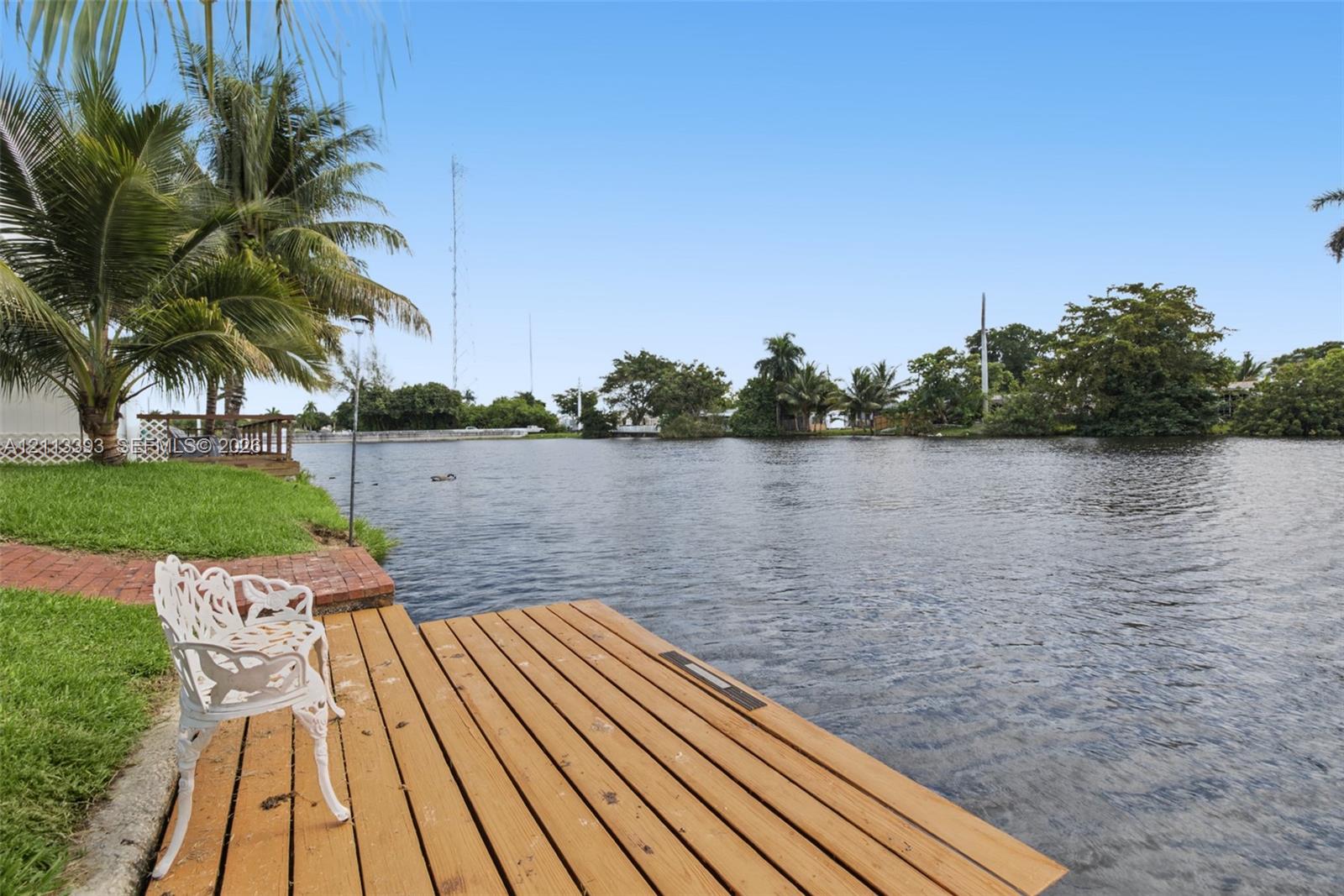 Waterfront lot featuring a wooden dock, brick paver pathway, and mature palm trees