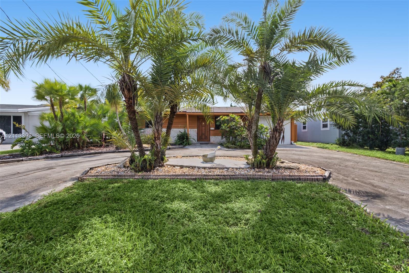 Exterior facade featuring wood-finish siding, a circular asphalt driveway, mature palm trees, and a central landscaped island with stone ground cover