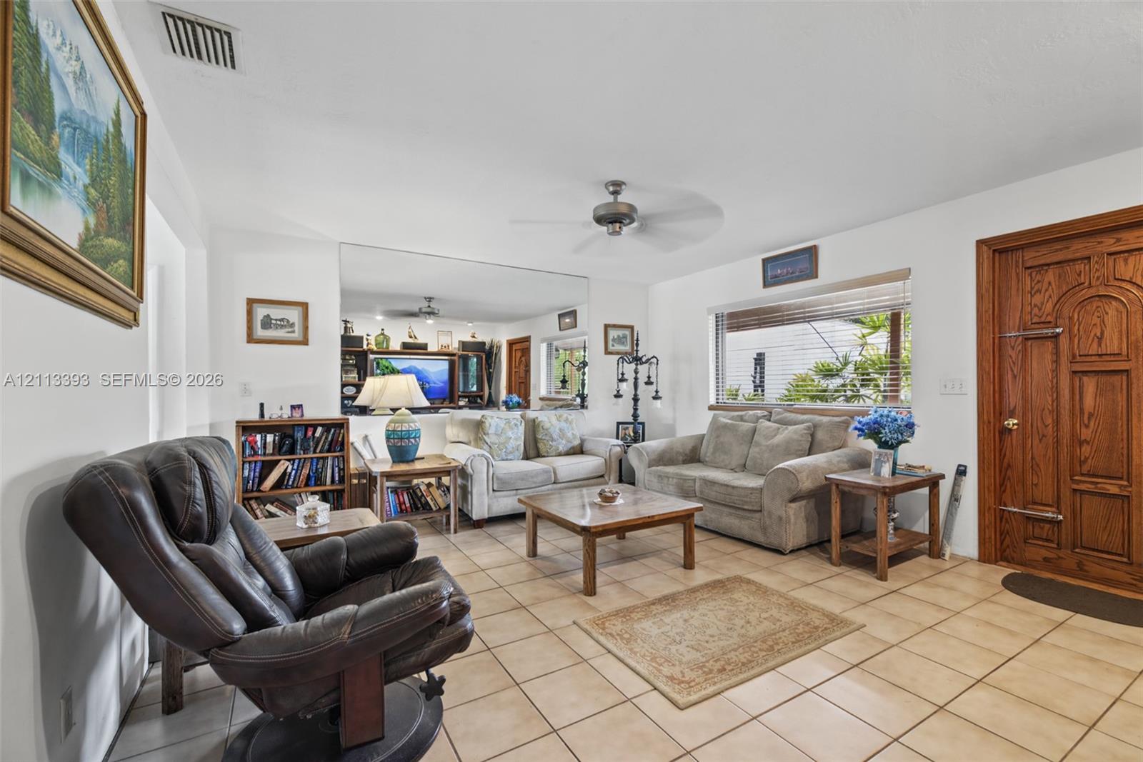 Living area featuring ceramic tile flooring, a large wall-mounted mirror, a ceiling fan, and a wood-finished entry door with ornate paneling