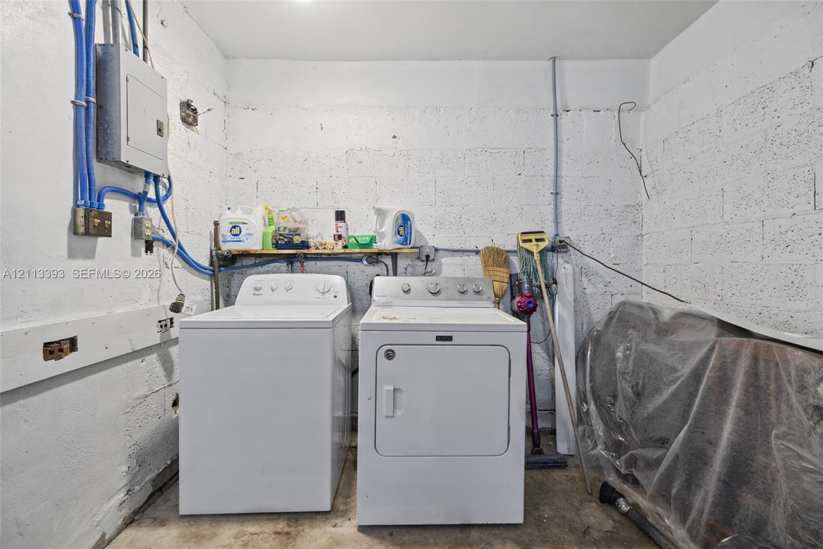 Utility area featuring exposed masonry walls, concrete flooring, built-in shelving, and dedicated laundry hookups with an electrical service panel