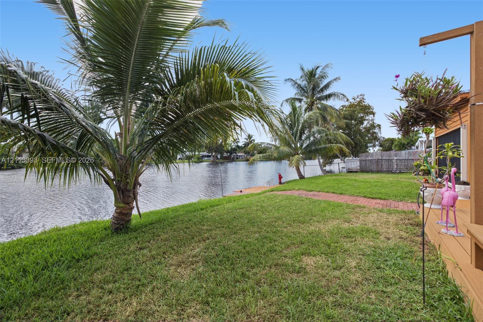 Waterfront backyard featuring a grass lawn, mature palm trees, a brick paver pathway, and an attached wood-finish deck