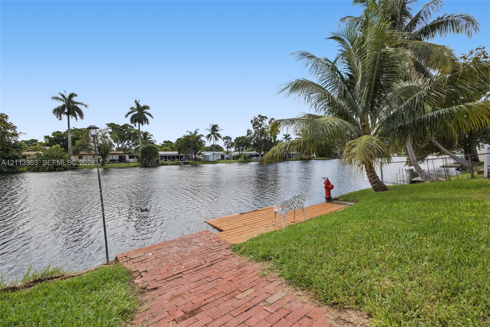 Waterfront property featuring a wood-plank dock, red brick path, manicured lawn, and mature palm trees