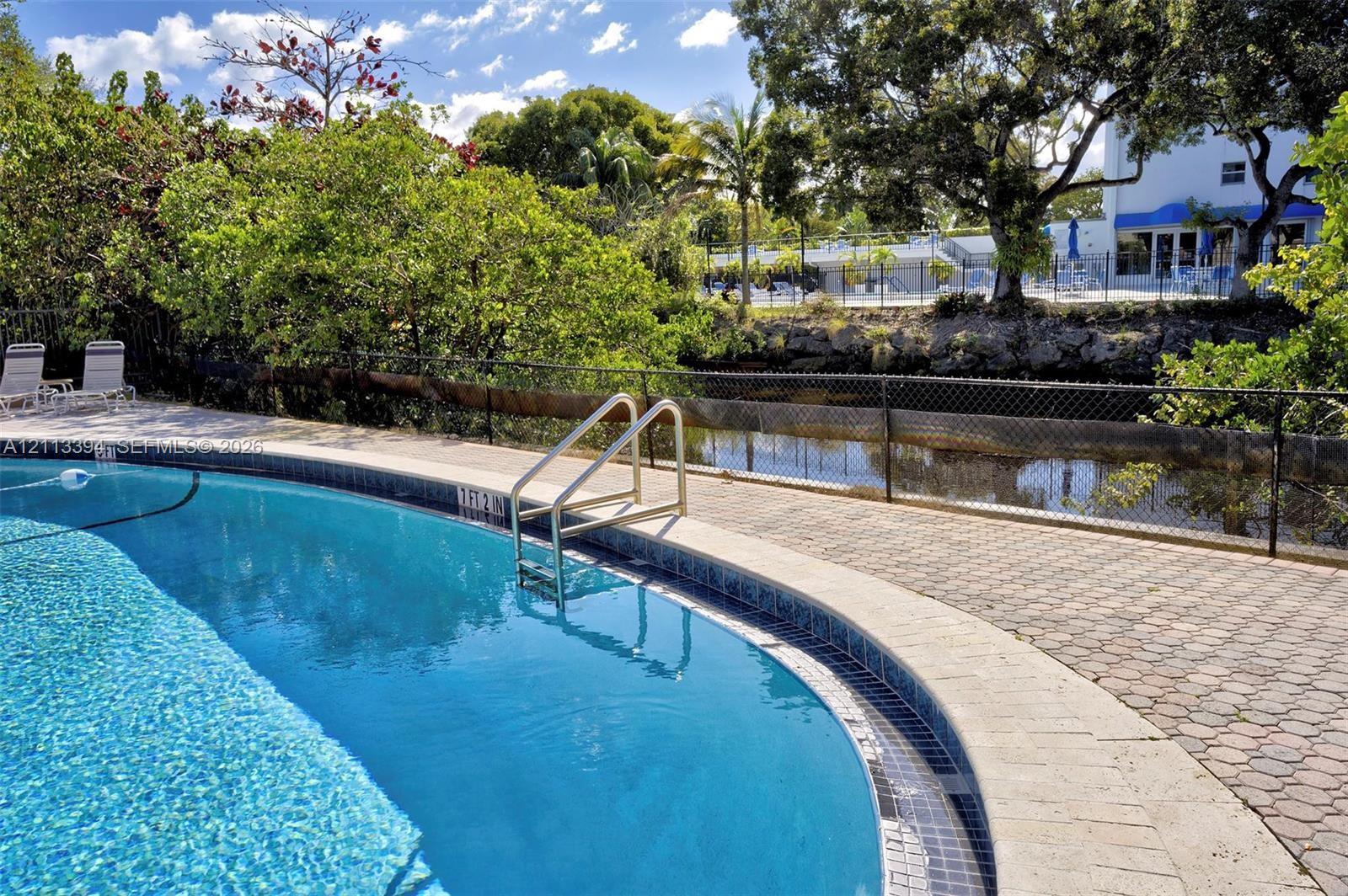 Curved in-ground swimming pool with metal handrails, stone-paver pool deck, and perimeter fencing