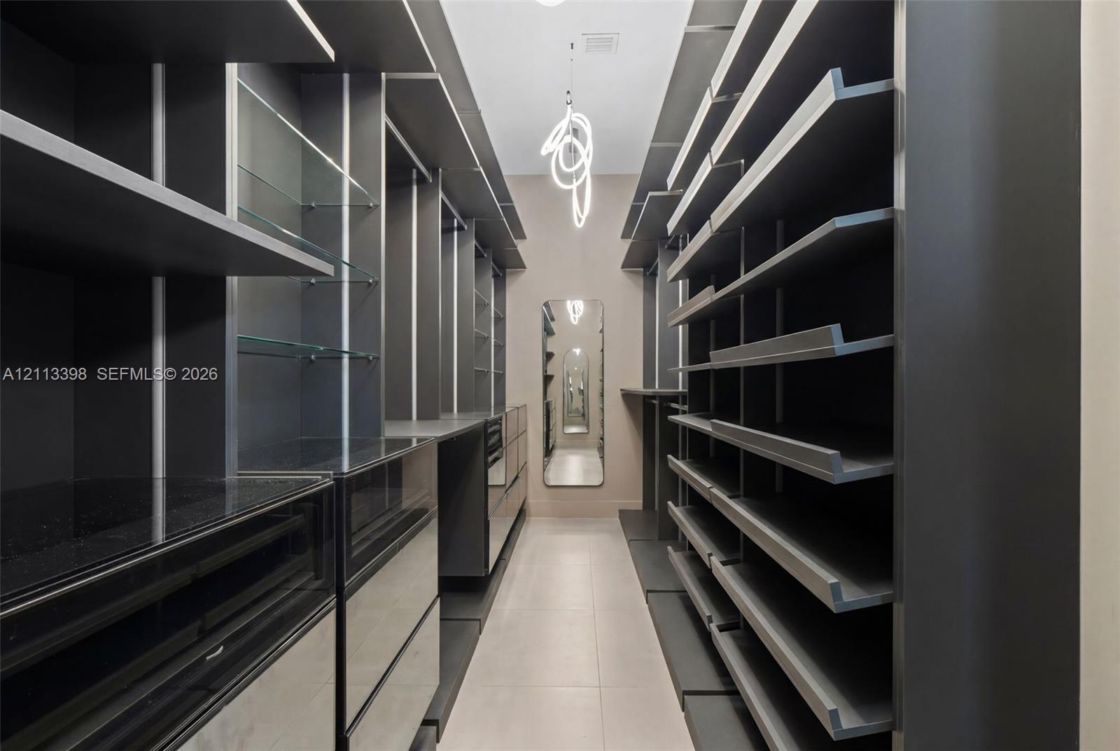Custom walk-in storage area featuring dark-toned built-in cabinetry, integrated glass shelving, sleek flat-front drawers, a full-length wall mirror, and a contemporary sculptural pendant fixture