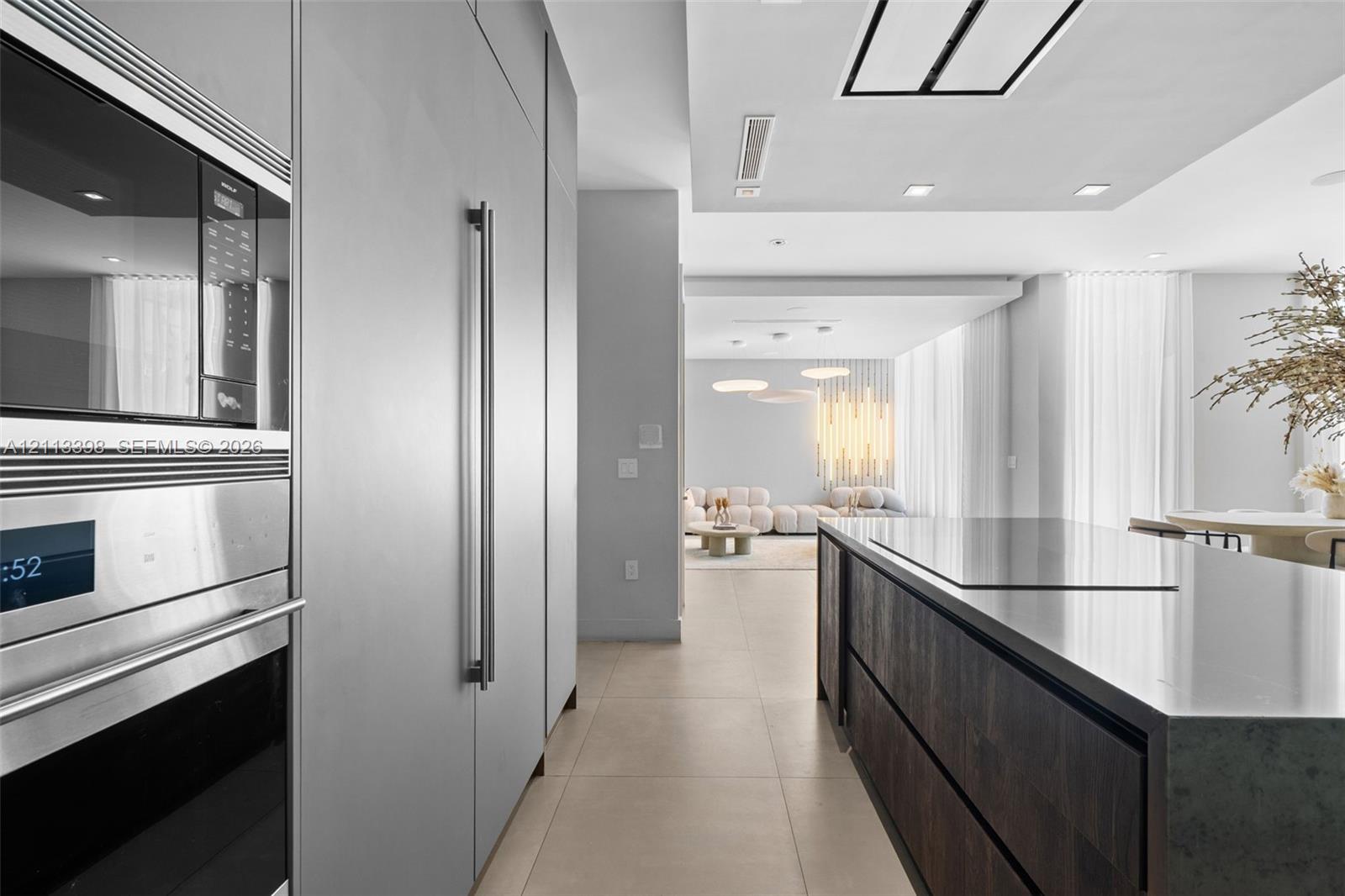 Kitchen featuring stainless steel built-in microwave and oven, floor-to-ceiling matte cabinetry, stone-surfaced kitchen island with integrated cooktop, wood-finish cabinetry, and neutral tile flooring