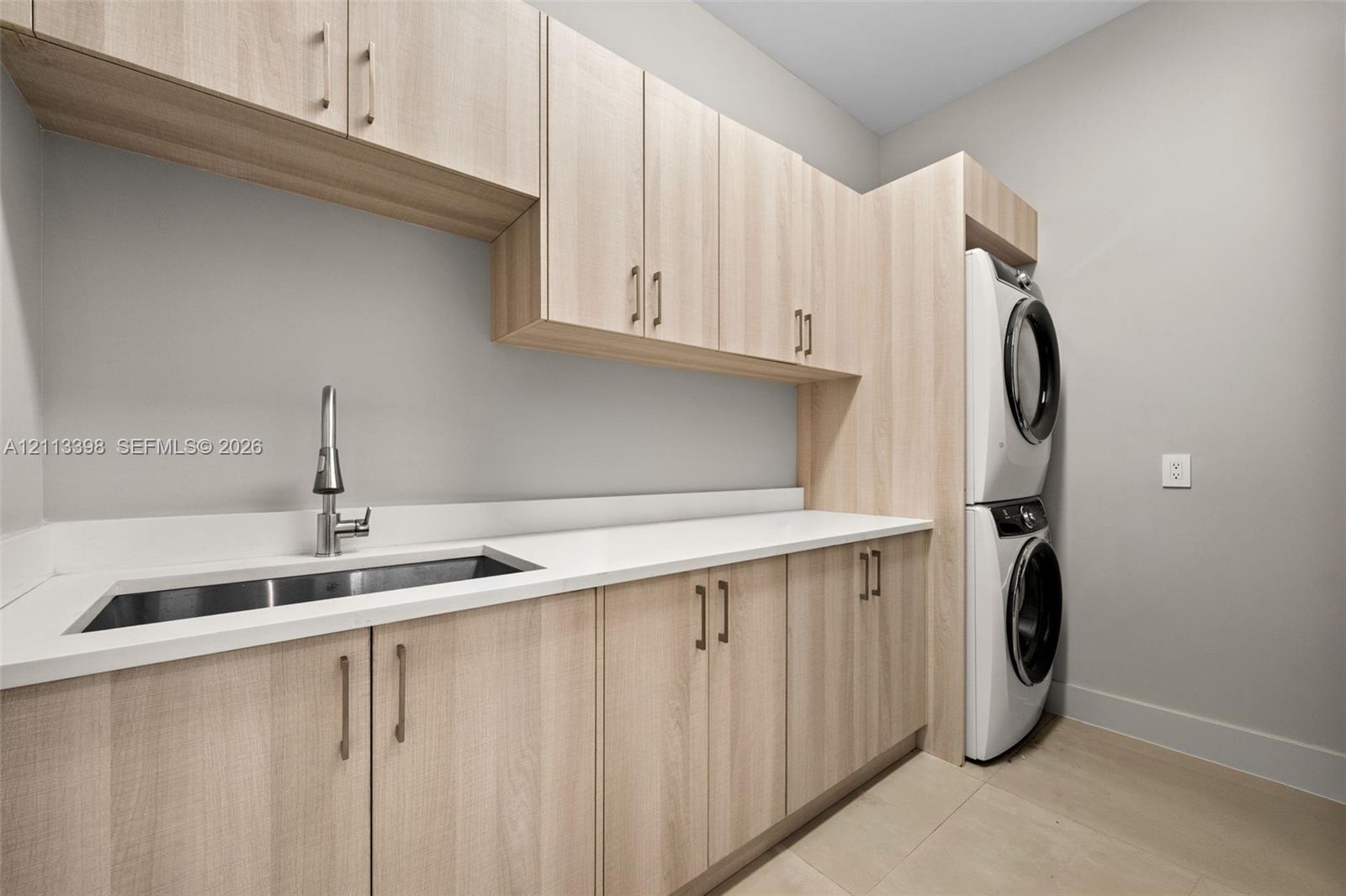 Laundry room featuring light wood-finish cabinetry, white solid-surface countertops, an undermount stainless steel sink with a high-arch pull-down faucet, and a stacked washer and dryer unit
