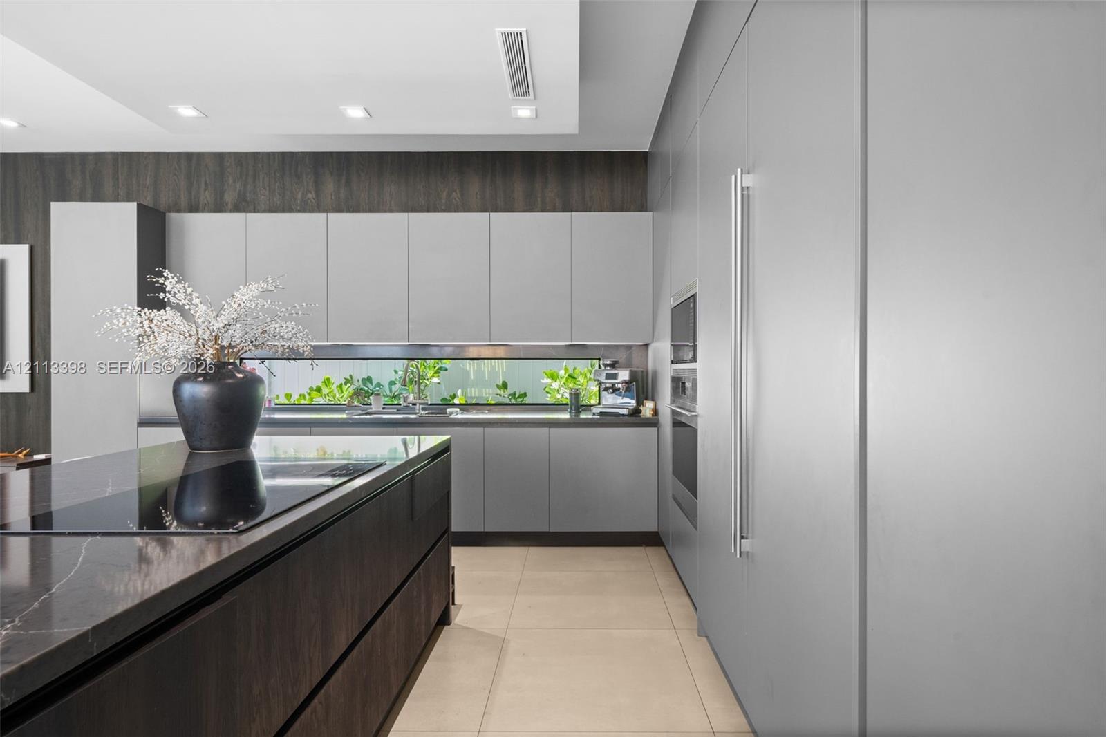 Contemporary kitchen featuring flat-panel cabinetry, a dark-toned center island with a stone-surface countertop, integrated stainless steel appliances, a linear backsplash window, and large-format tile flooring