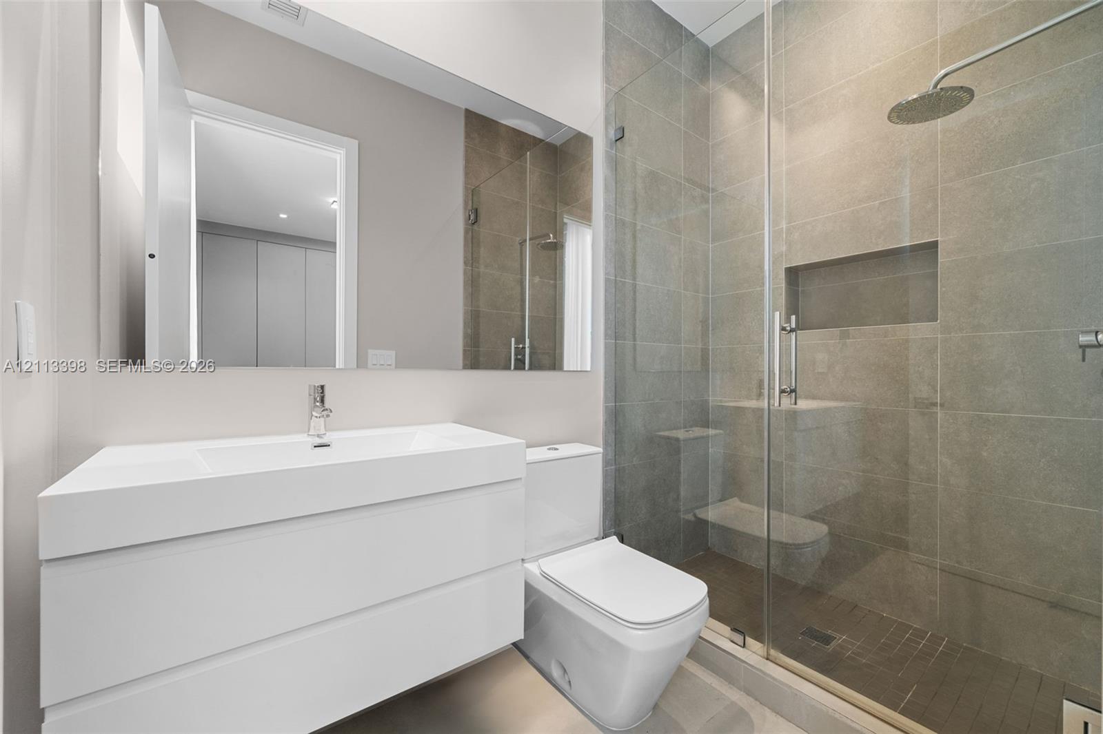 Modern wall-mounted vanity with integrated sink, full-width mirror, walk-in shower with floor-to-ceiling stone-finish tile, glass shower enclosure, and built-in shower niche