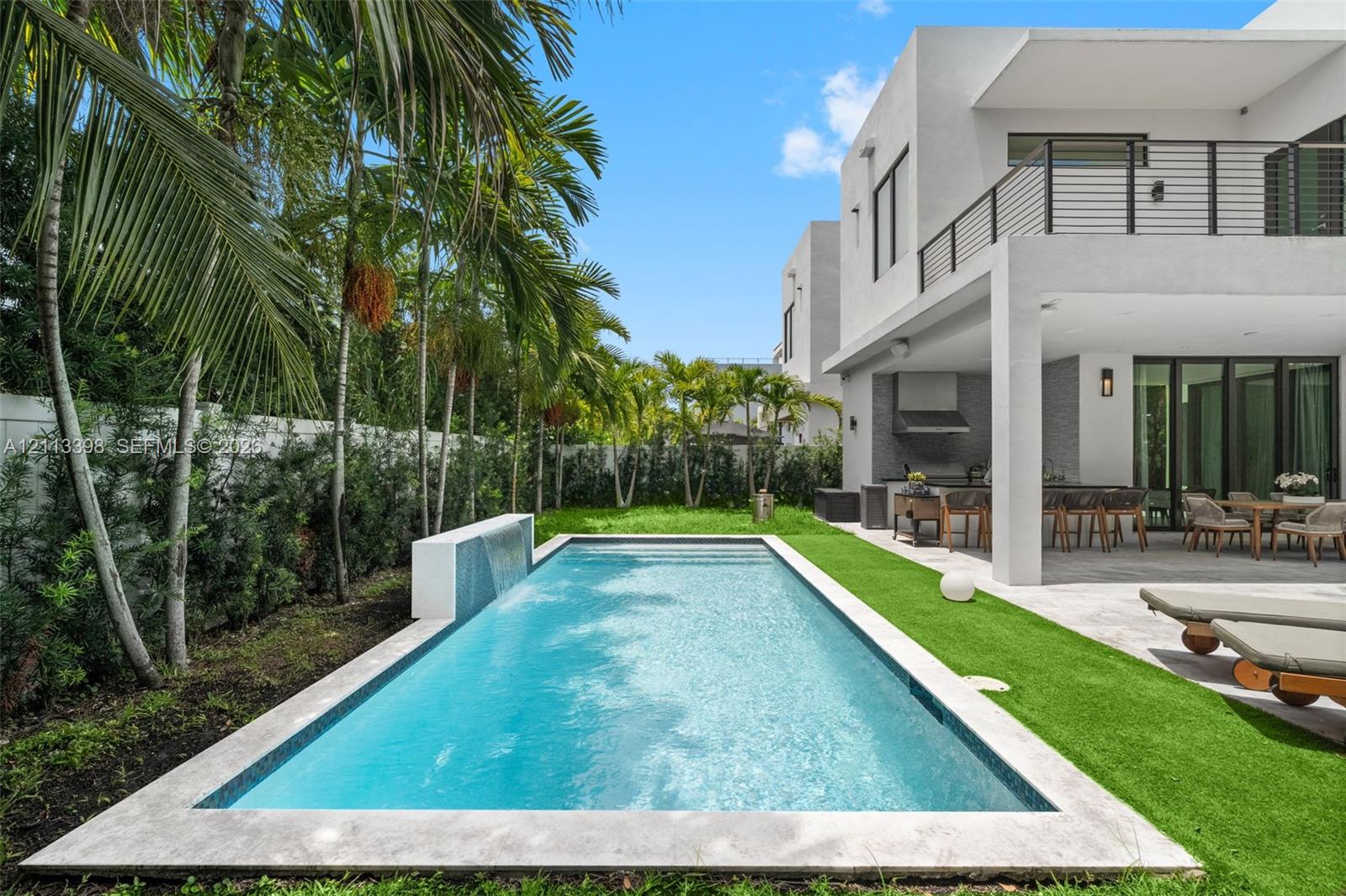 Rear exterior featuring a rectangular pool with a built-in water feature, natural stone coping, covered patio, outdoor kitchen, and synthetic turf lawn