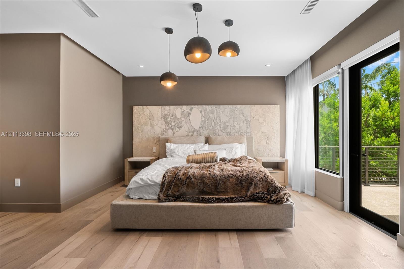 Custom full-height stone-textured headboard wall, light-toned wood-finish flooring, trio of suspended pendant lights, and floor-to-ceiling glass sliding doors opening to a balcony