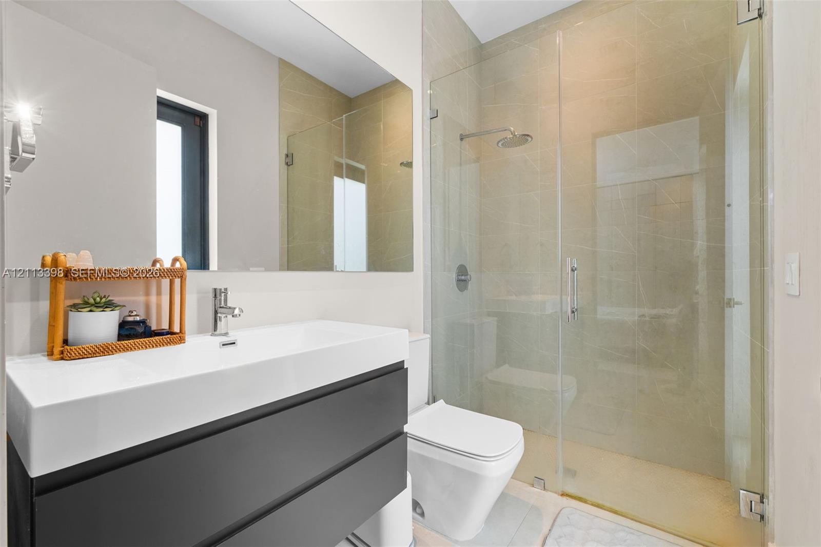 Wall-mounted dark cabinetry with a white rectangular sink basin, large frameless wall mirror, walk-in glass-enclosed shower, and neutral-toned wall tiling