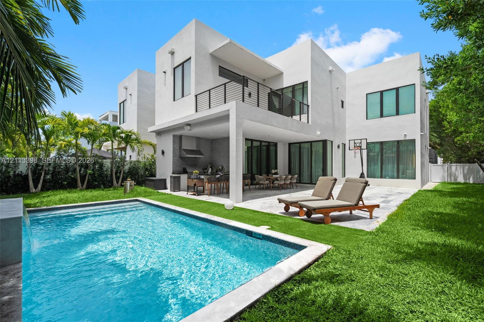 Contemporary residence featuring a stucco exterior, second-story balcony with metal railing, covered outdoor kitchen, stone-paved patio, and a swimming pool with a stone water feature
