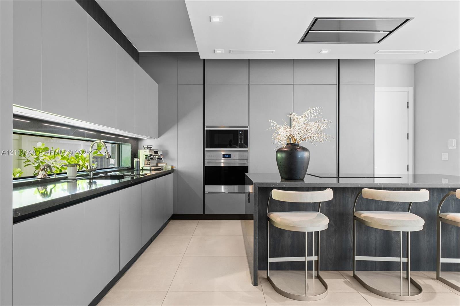 Modern kitchen featuring matte grey flat-panel cabinetry, dark stone countertops, built-in stainless steel appliances, a horizontal window backsplash, and a ceiling-mounted ventilation hood