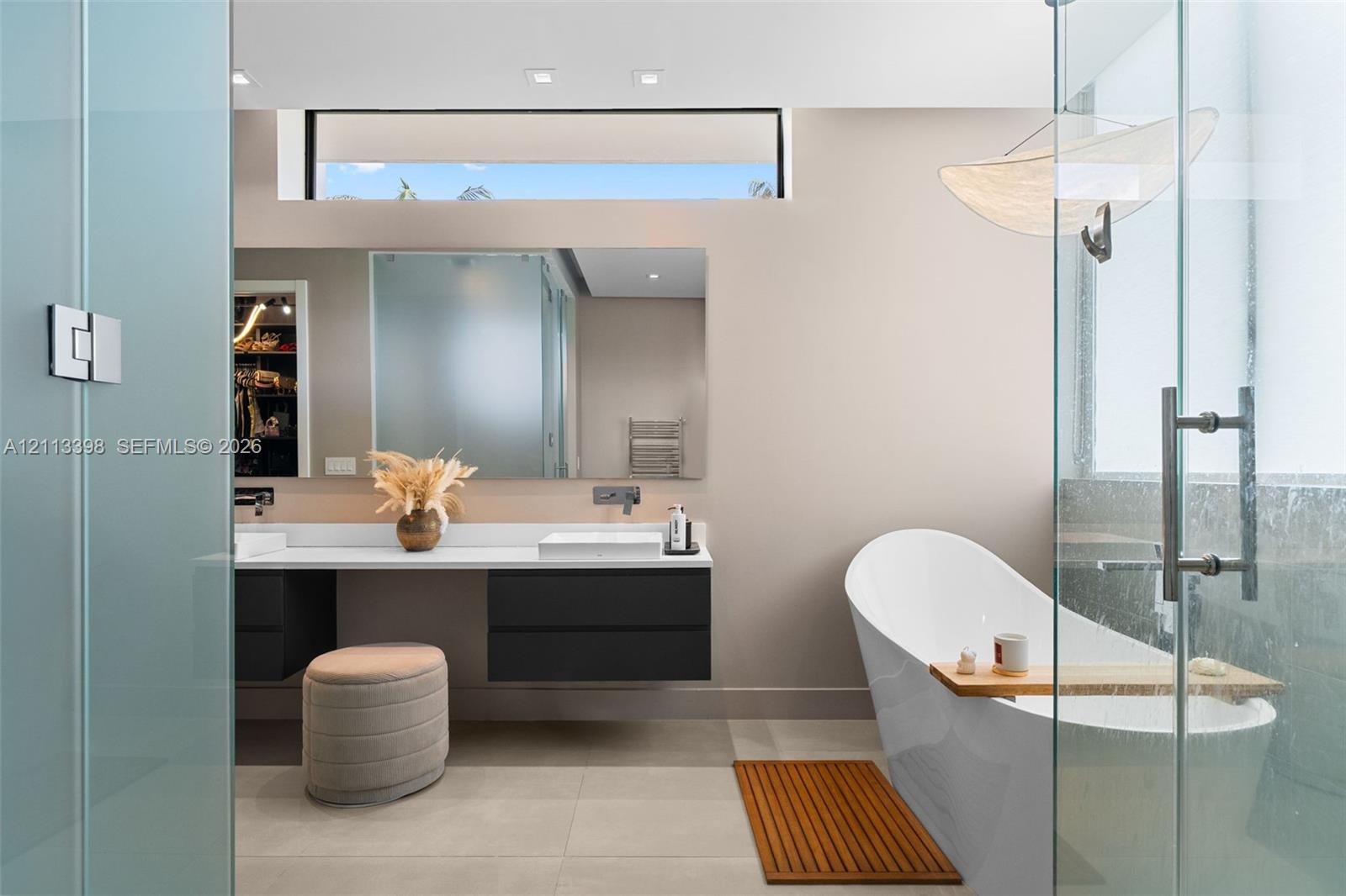 Floating dual-vanity with matte cabinetry and solid-surface countertop, freestanding soaking tub, frameless glass-enclosed shower, clerestory window, and large-format tile flooring