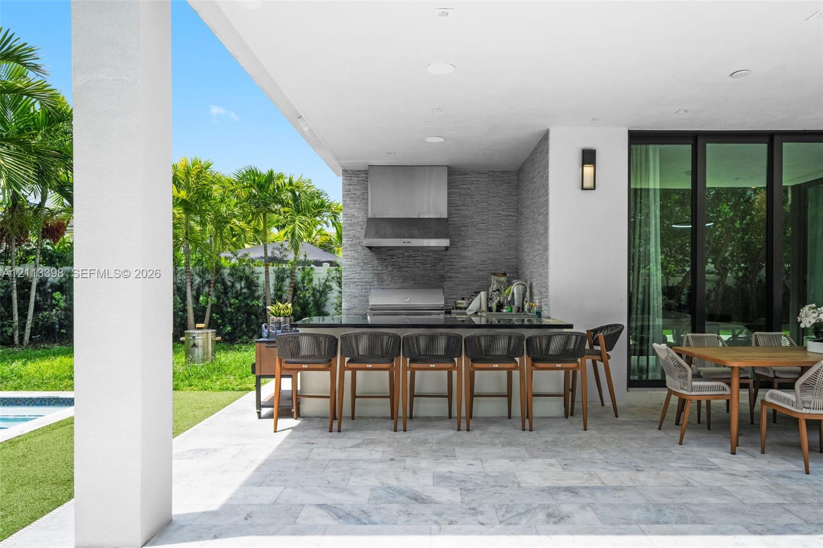 Covered outdoor patio featuring light-toned stone tile flooring, a built-in outdoor kitchen with a stainless steel range hood, natural stone backsplash, and dark stone countertop