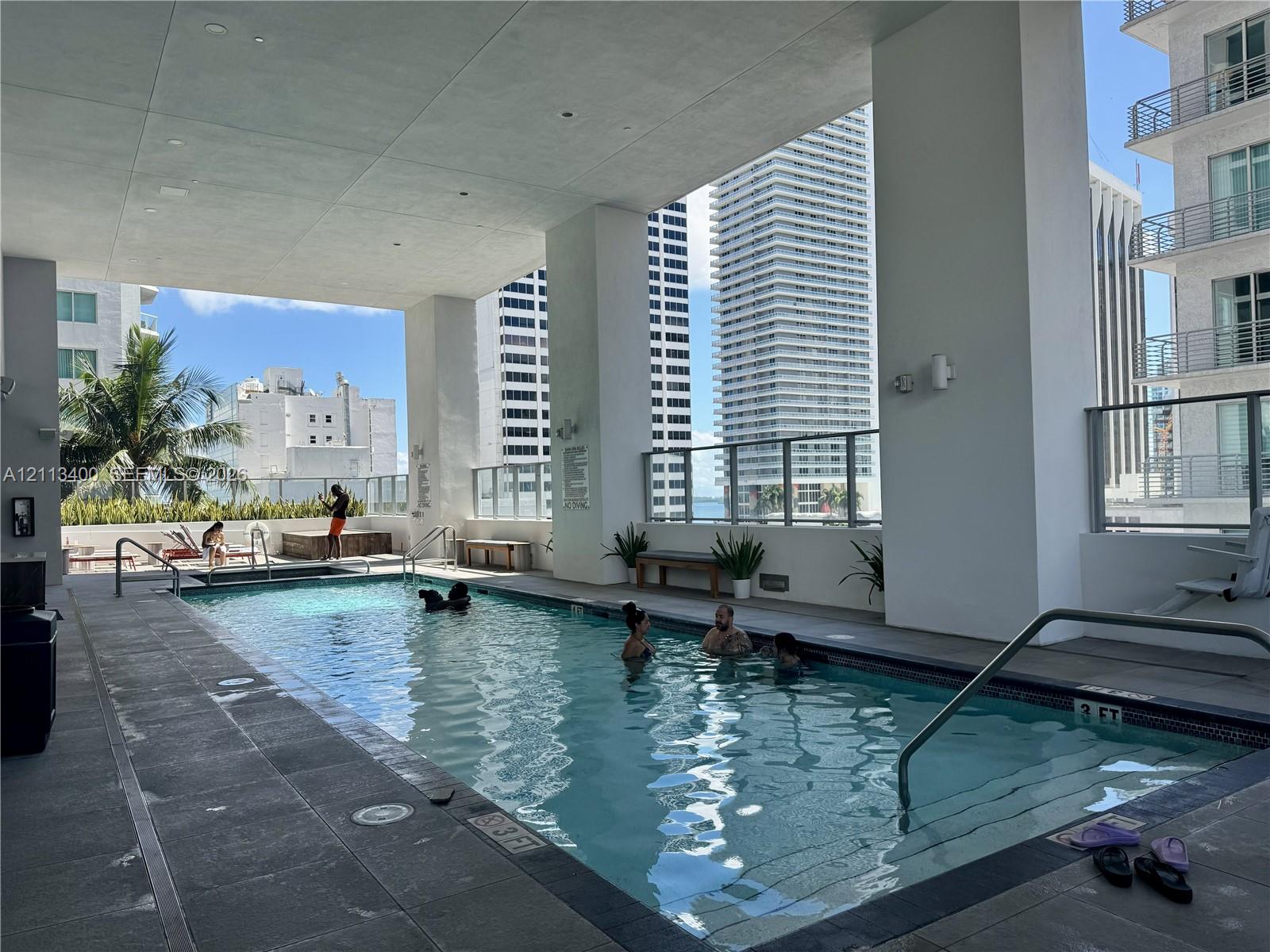 Covered outdoor swimming pool featuring concrete decking, metal railings, and a tiled pool edge Covered outdoor swimming pool featuring concrete decking, metal railings, and a tiled pool edge