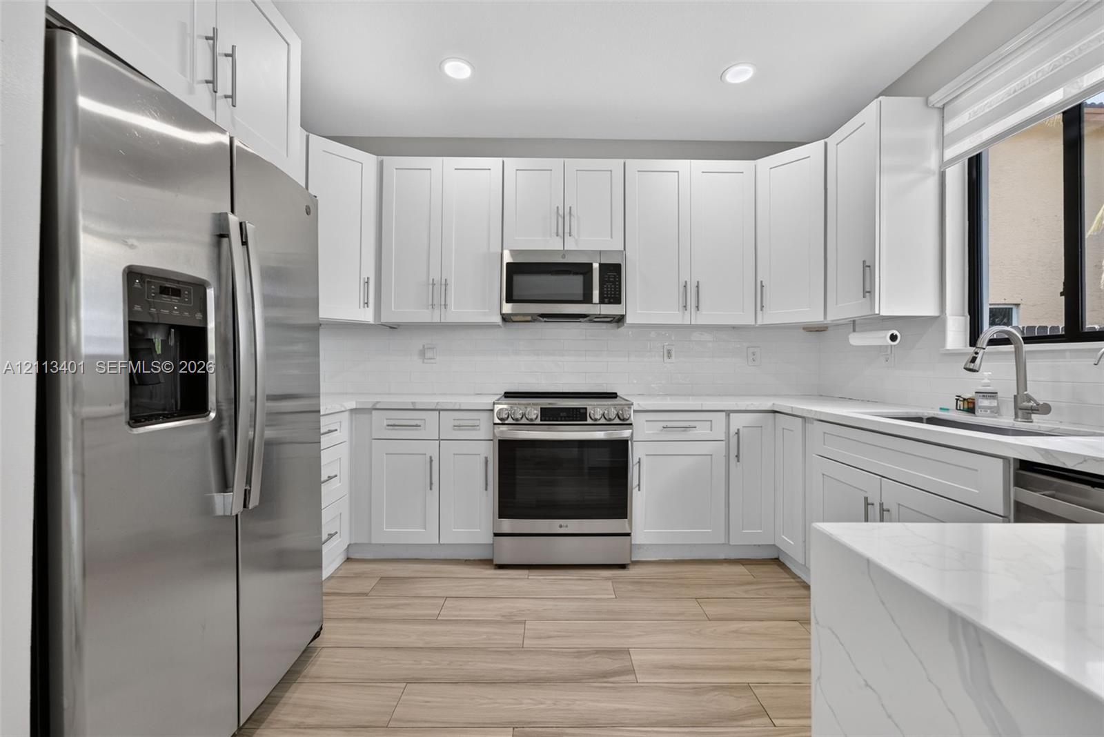 Kitchen featuring white Shaker-style cabinetry, stainless steel appliances, quartz countertops with a subtle veining pattern, white subway tile backsplash, and wood-finish flooring