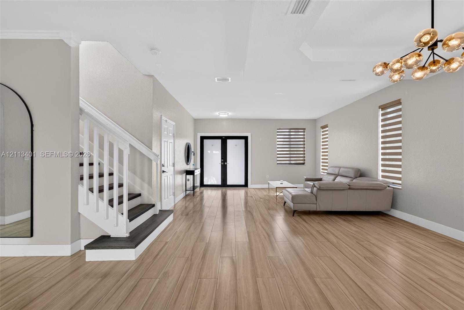 Wood-finish flooring, white staircase with dark treads, tray ceiling, multi-bulb chandelier, and glass-paneled double entry doors