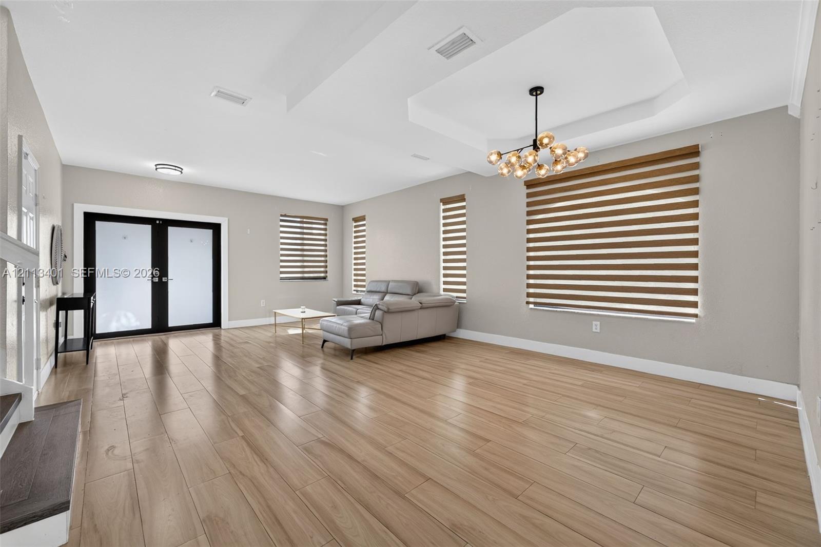 Open-concept room featuring tray ceilings, wood-finish flooring, a multi-globe chandelier, double-entry glass doors, and layered window shades