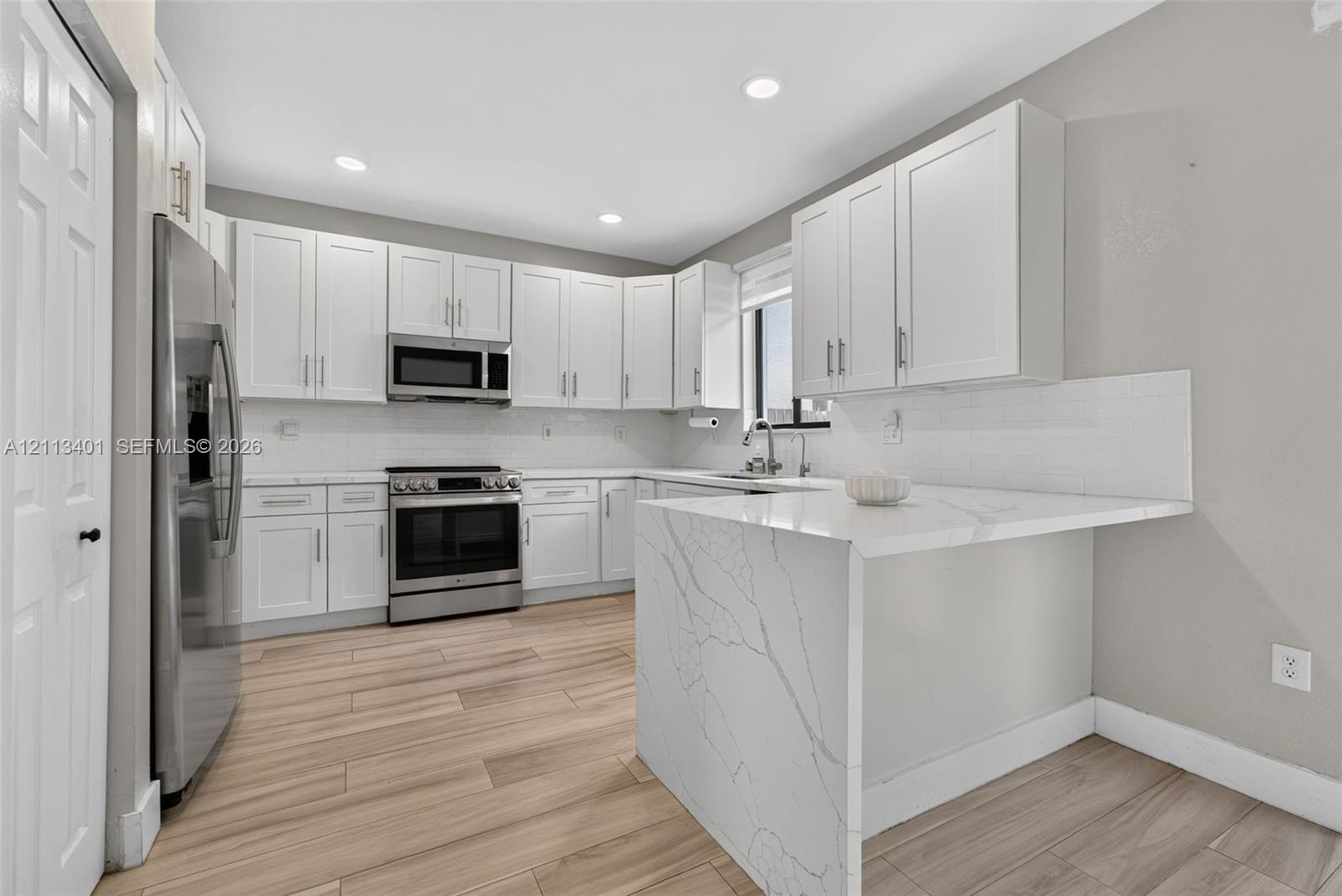 Kitchen featuring white shaker-style cabinetry, a stone-patterned waterfall countertop, stainless steel appliances, and wood-finish flooring
