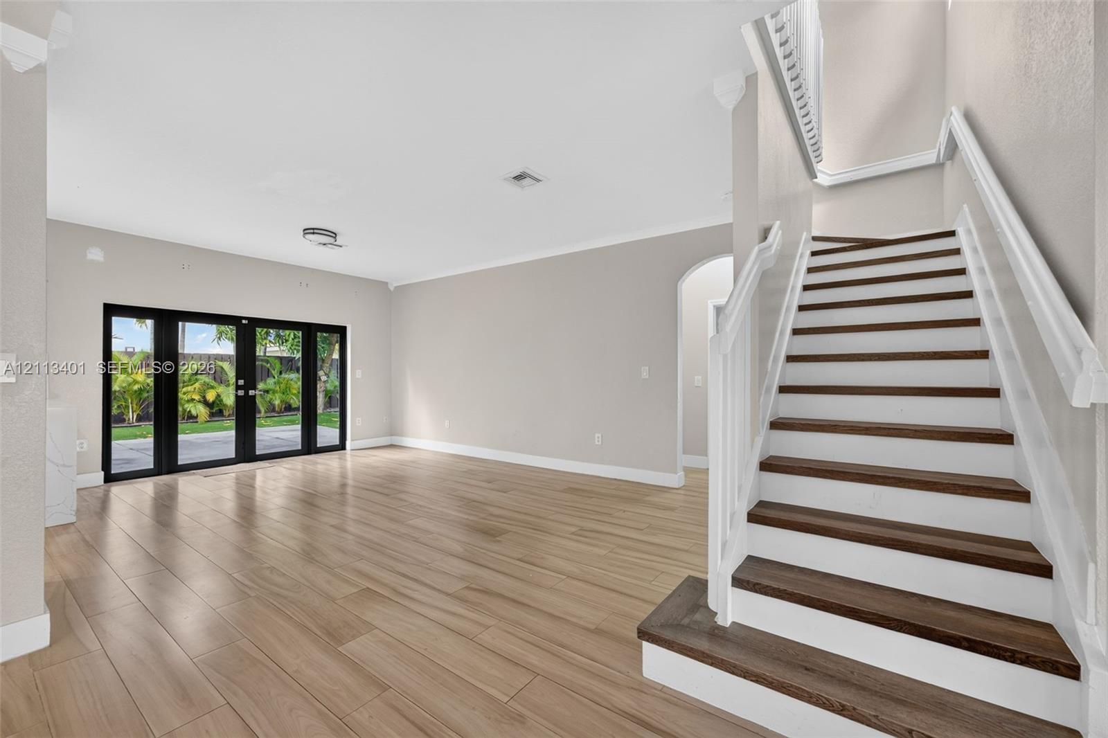 Open-concept interior featuring wood-finish flooring, a staircase with white balusters and dark wood-finish treads, crown molding, and black-framed glass French doors