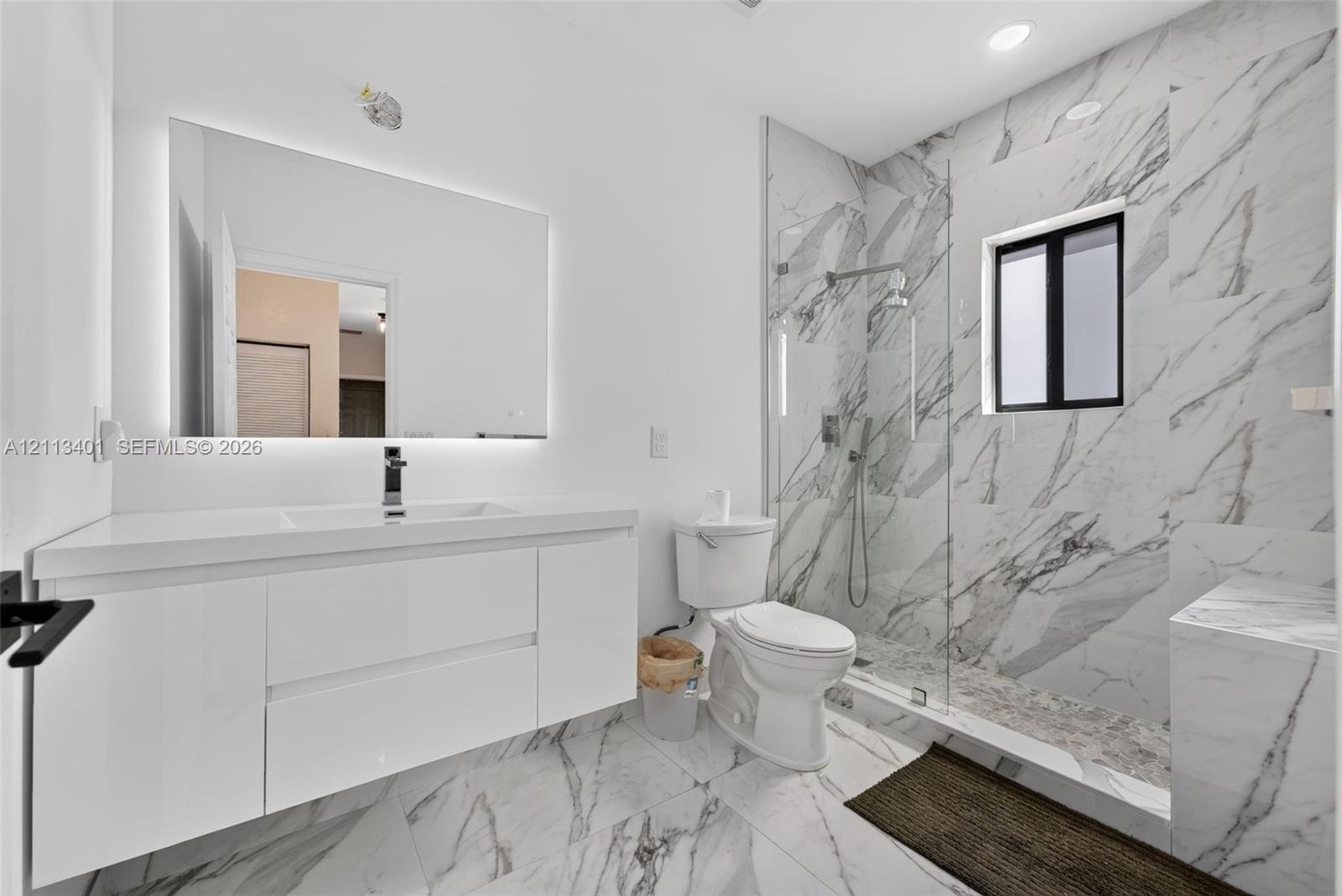 Floating white vanity with integrated sink, expansive LED-backlit mirror, floor-to-ceiling stone-look wall tiles, walk-in glass-enclosed shower, and matching stone-look tile flooring