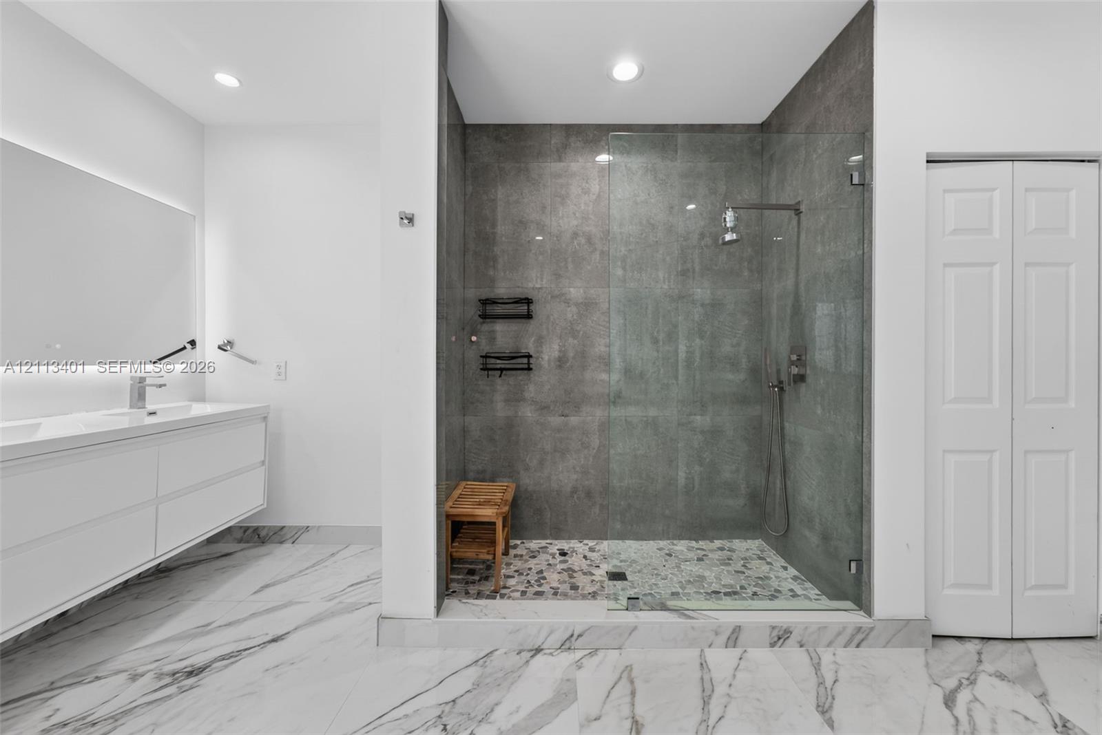 Floating dual-sink vanity with under-cabinet lighting, walk-in shower with frameless glass enclosure, floor-to-ceiling stone-look wall tile, pebble-texture shower flooring, and large-format porcelain tile flooring