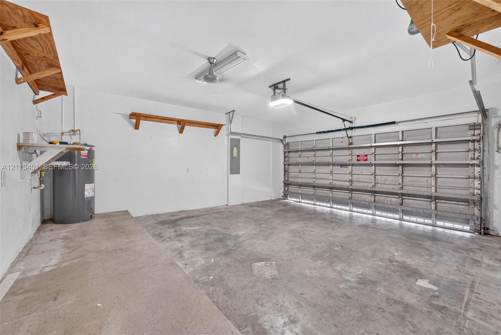 Two-car garage featuring a concrete floor, built-in wooden wall shelving, overhead fluorescent lighting, an automated door opener, and a dedicated water heater