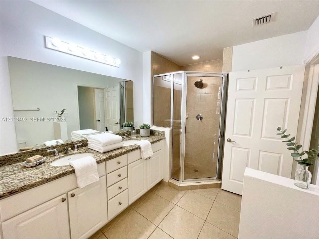 Bathroom featuring a dual-sink vanity with stone-surface countertops, expansive wall-mounted mirror, corner glass-enclosed shower, and tile flooring