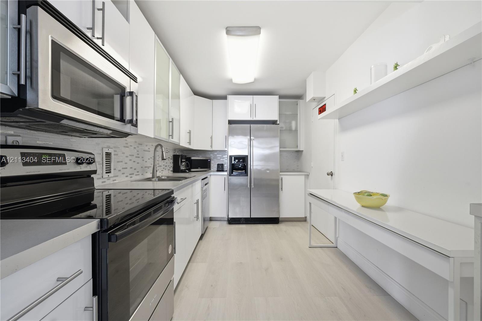 Galley kitchen featuring high-gloss white cabinetry, stainless steel appliances, light-toned wood-finish flooring, a decorative tile backsplash, and an integrated wall-mounted counter surface with matching shelving