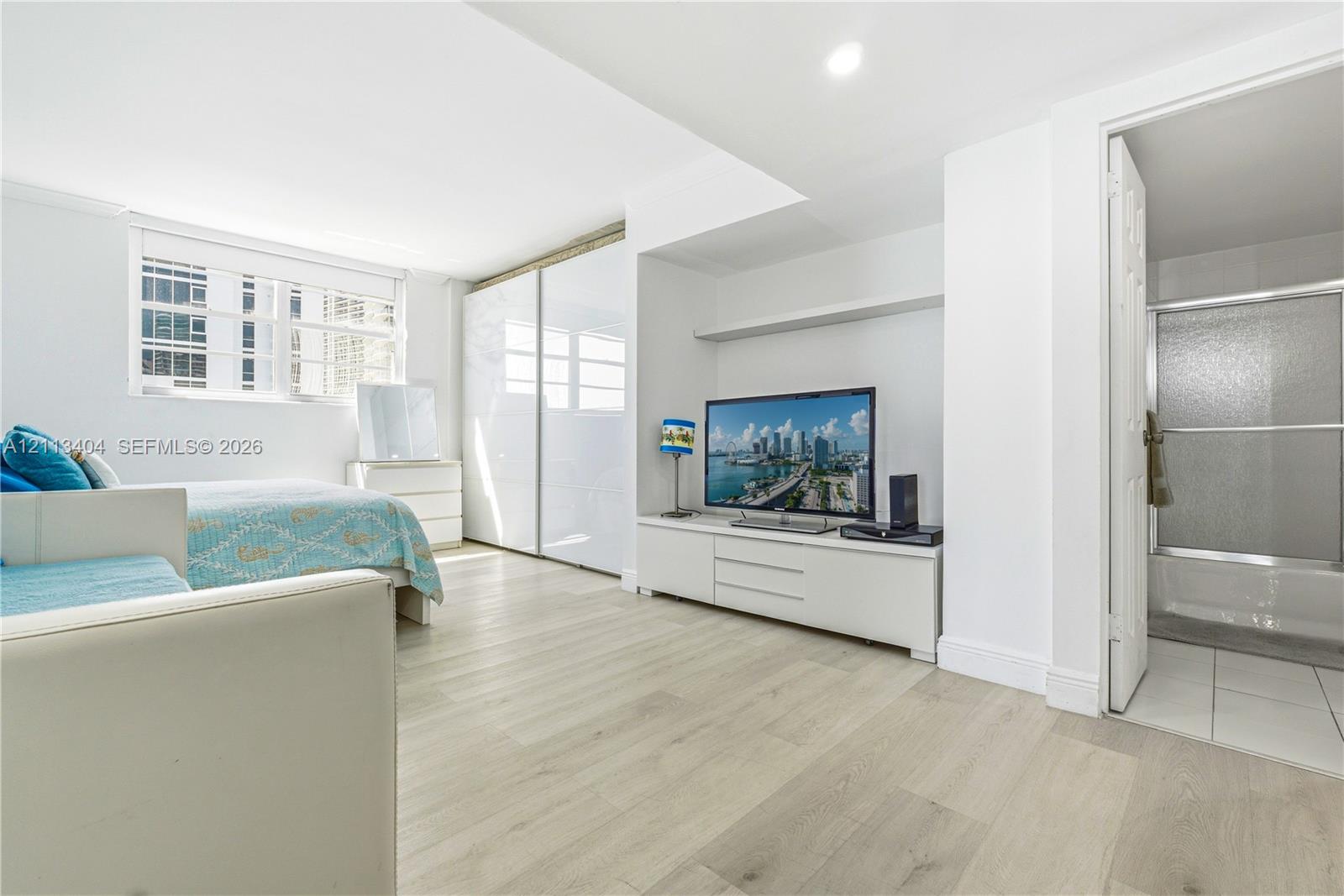 Light-filled room featuring wood-finish flooring, sliding-door closets with high-gloss panels, recessed lighting, and a bathroom containing a shower-tub combination
