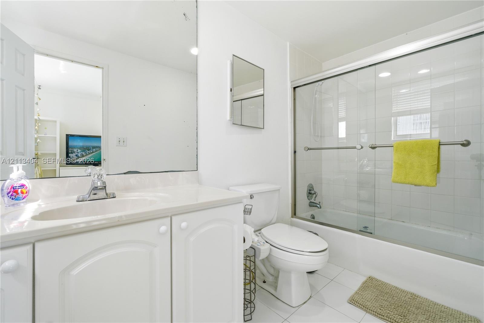 Bathroom featuring a white vanity cabinet, integrated sink with a chrome faucet, large wall-mounted mirror, white ceramic tile flooring, and a bathtub with sliding glass doors and white tile surround