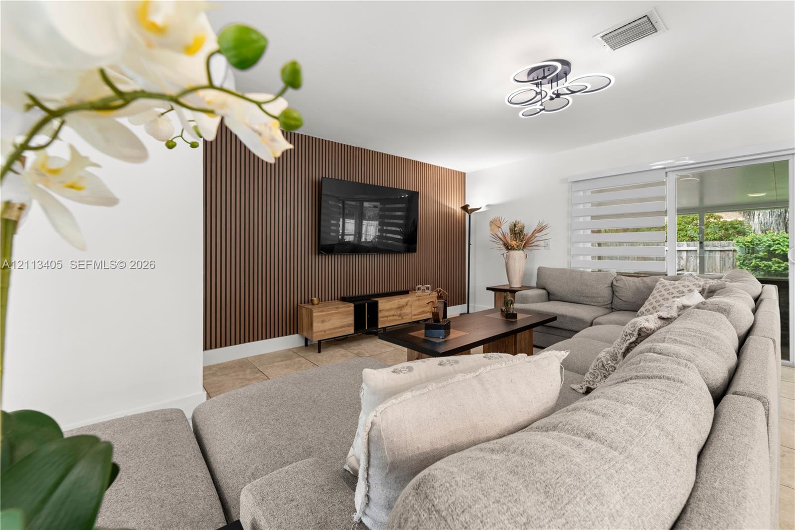 Living space featuring a vertical slat wood-finish accent wall, tile flooring, modern multi-ring ceiling light fixture, and large sliding glass door with horizontal blinds