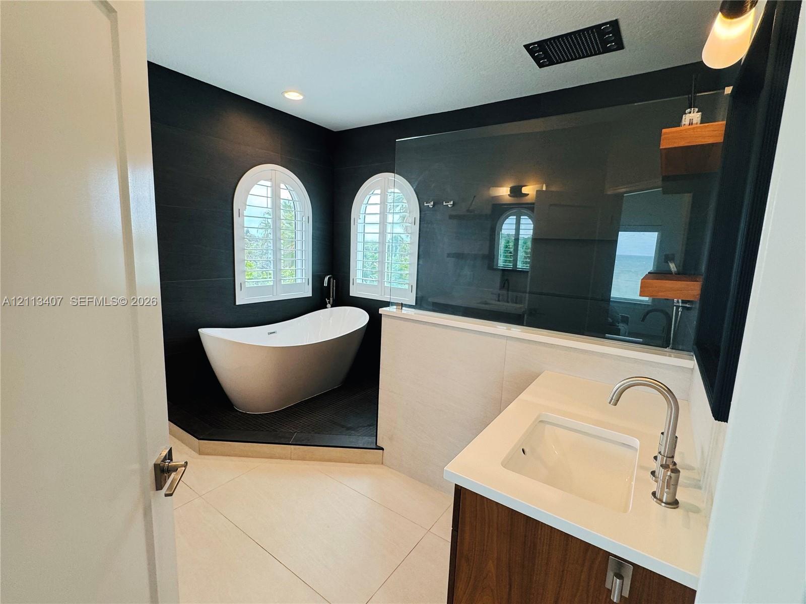 Second floor Bathroom featuring a freestanding soaking tub, arched windows with shutters, dark tile accent walls, a vanity with a white countertop and rectangular sink, and floating wood shelving