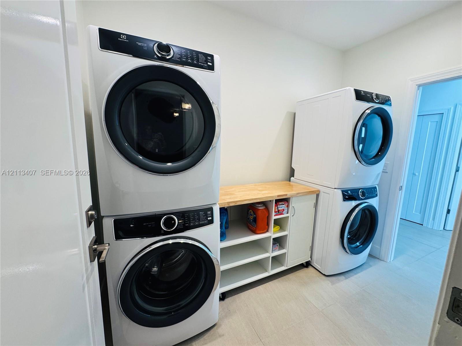 Laundry room featuring dual sets of stackable front-loading washer and dryer units, white custom cabinetry with open shelving, and a natural wood countertop Laundry room featuring dual sets of stackable front-loading washer and dryer units, white custom cabinetry with open shelving, and a natural wood countertop