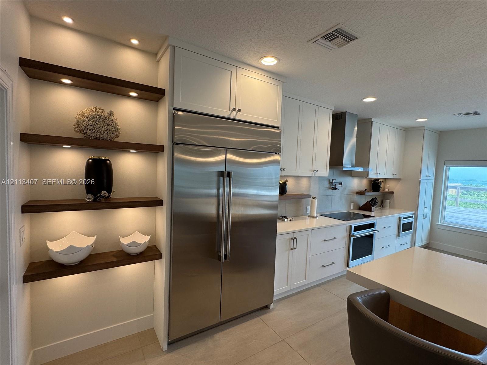 Kitchen featuring recessed lighting, stainless steel refrigerator, white cabinetry, solid-surface countertops, and built-in wood shelving Kitchen featuring recessed lighting, stainless steel refrigerator, white cabinetry, solid-surface countertops, and built-in wood shelving