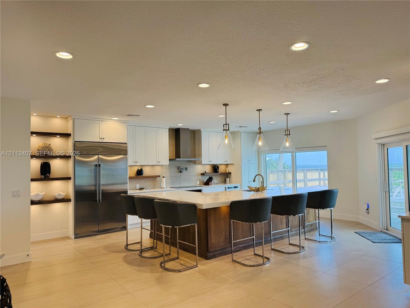 Kitchen featuring a large center island with stone countertop, stainless steel refrigerator, recessed lighting, glass pendant fixtures, and built-in shelving with integrated lighting Kitchen featuring a large center island with stone countertop, stainless steel refrigerator, recessed lighting, glass pendant fixtures, and built-in shelving with integrated lighting
