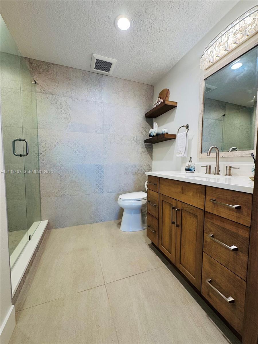 Bathroom featuring a wood-finish vanity with a light-toned countertop, floor-to-ceiling patterned tile shower wall, glass-enclosed shower, dual-tone wall-mounted shelving, and recessed lighting Bathroom featuring a wood-finish vanity with a light-toned countertop, floor-to-ceiling patterned tile shower wall, glass-enclosed shower, dual-tone wall-mounted shelving, and recessed lighting