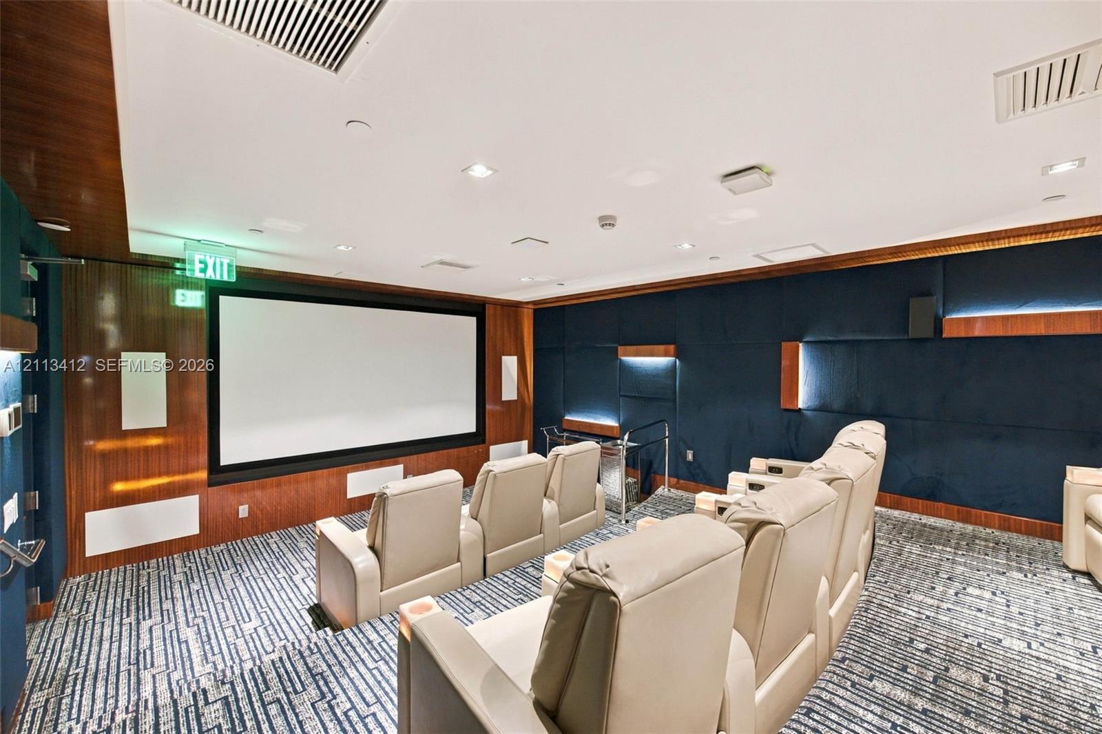 Media room featuring tiered flooring, dark blue upholstered wall panels, integrated wood-finish wall accents, recessed lighting, and a built-in projection screen
