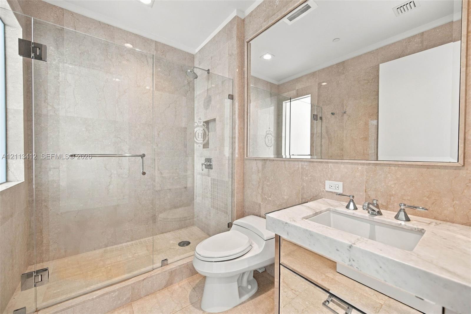 Full-wall stone tiling, glass-enclosed walk-in shower with built-in recessed niche, vanity with integrated basin, polished stone countertop, and large framed mirror