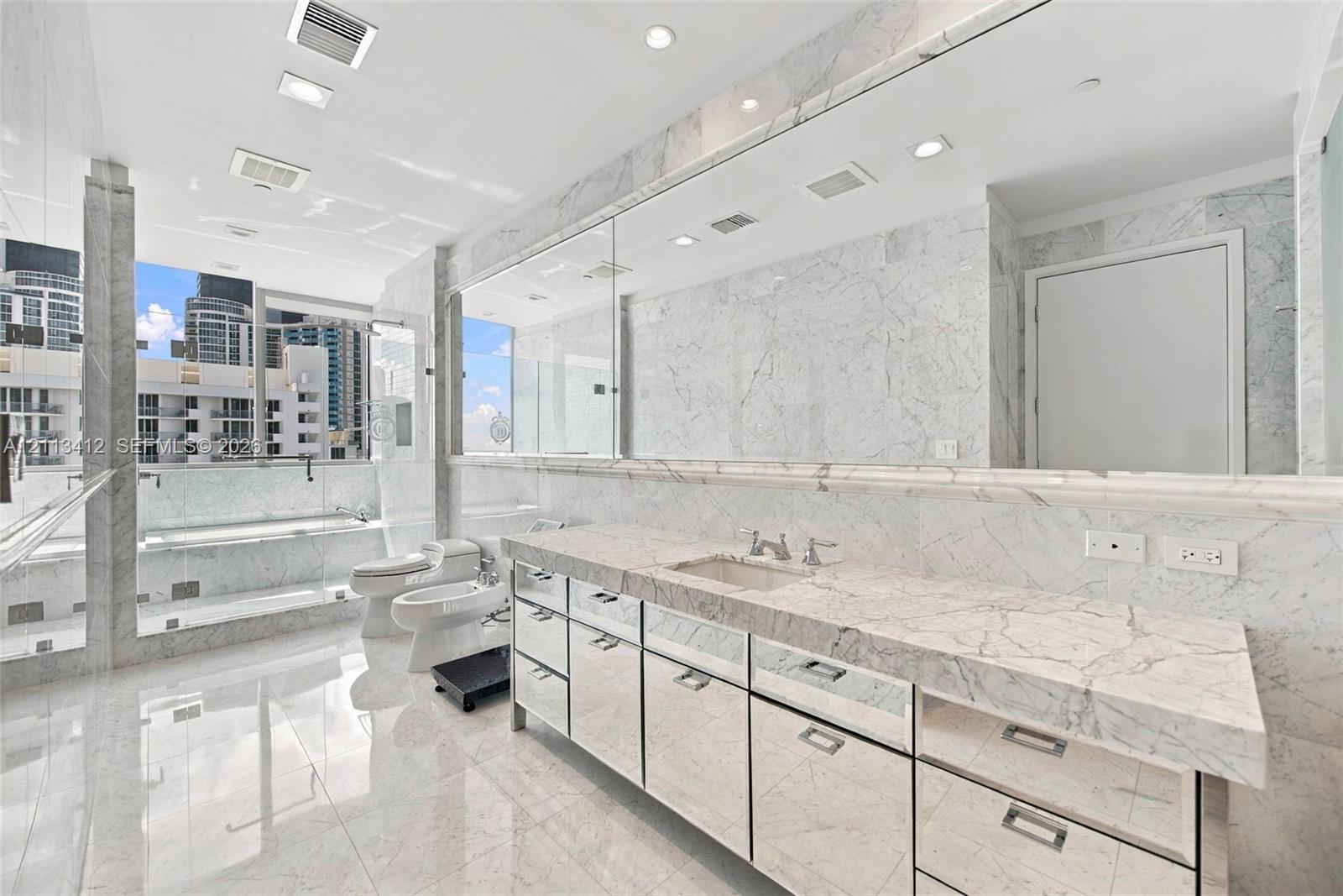 Full-length stone vanity with dual undermount sinks, expansive wall-to-wall mirror, floor-to-ceiling natural stone wall and floor tile, glass-enclosed shower, and built-in vanity cabinetry